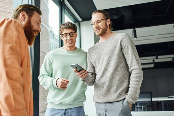 businessman in eyeglasses and stylish casual clothes standing with hand in pocket and networking on mobile phone near smiling colleagues in contemporary office, concept of successful collaboration