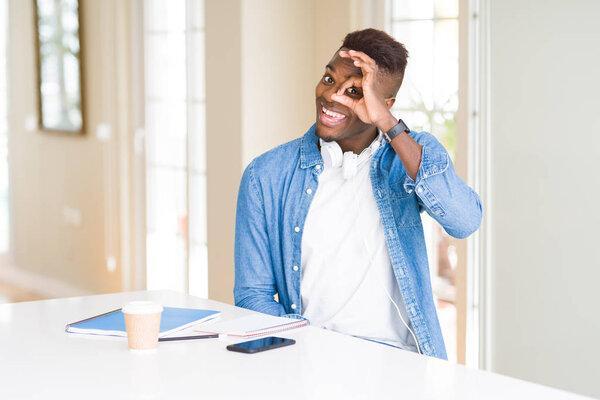 African american student man studying using notebooks and wearing headphones doing ok gesture with hand smiling, eye looking through fingers with happy face.