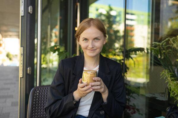 Close up of young female in business casual wear drinking cup of coffee while sitting at small table and enjoying time in street cafe. High quality photo