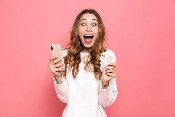 Portrait of a happy young woman using mobile phone, holding plastic credit card isolated over pink background