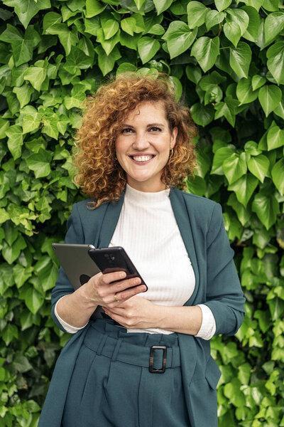 Happy business woman with curly hair and beautiful suit holding her phone and laptop and looking at camera.