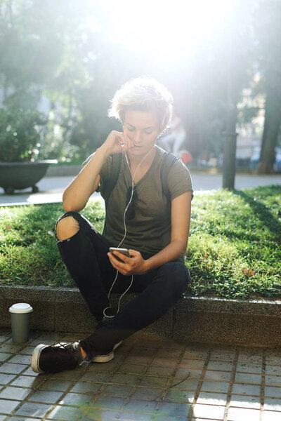 Blonde caucasian female wearing casual clothes is reading emails on the mobile phone while enjoying morning in a city park. A blank paper cup of takeaway coffee or tea is standing beside the girl.