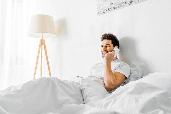 cheerful man talking on smartphone while sitting on bed in the morning