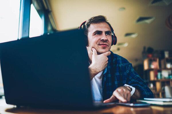 Young good looking positive male person enjoying recreation with big modern headphones and modern device indoors.Handsome happy hipster guy listening to favorite music during free time in cafe