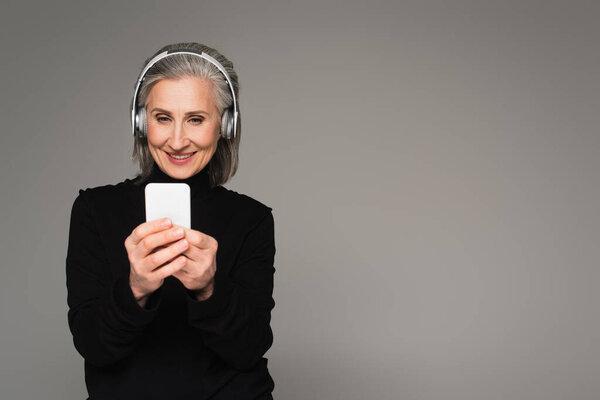 Middle aged woman in headphones using smartphone isolated on grey 