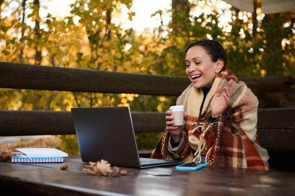 Cheerful middle-aged pretty woman warms herself wrapped in plaid blanket and drinking hot coffee from paper cup during video call while sitting on open country terrace on cool autumn day in oak grove