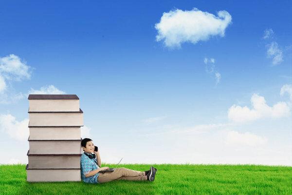 Picture of handsome preteen schoolboy speaking on a smartphone while sitting near a pile of books. Shot in the meadow