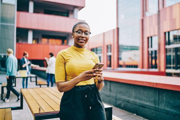 Positive african american woman in casual wear and spectacles looking at camera and holding mobile phone, portrait of attractive dark skinned hipster guy  using smartphone and 4G internet for chattin
