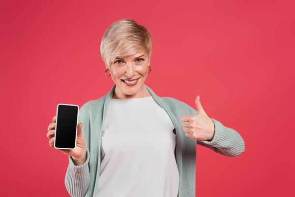 cheerful woman showing thumb up while holding cellphone with blank screen isolated on pink