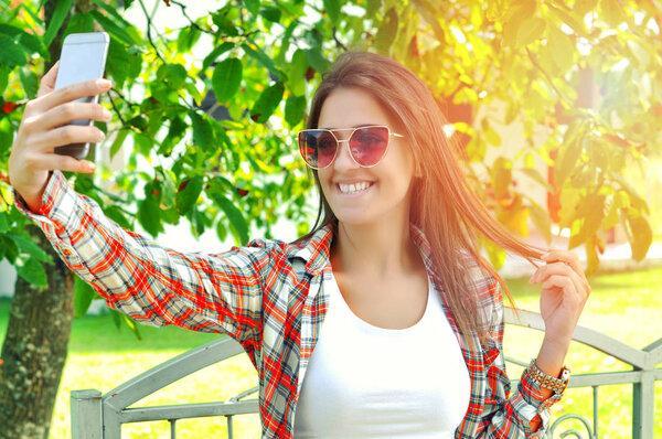  Beautiful smiling young woman making self-portrait with smartphone wearing casual checkered shirt sunglasses on sunny day on green nature background