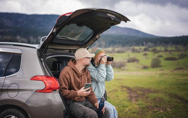 Romantic middle age caucasian couple traveling by car sitting in open trunk with photo camera and smartphone