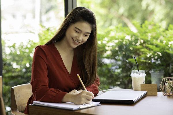 Beautiful Attractive Asian woman working with laptop smile and writing idea and requirement for success job outside office in coffee shop,Business Concept,Warm Tone