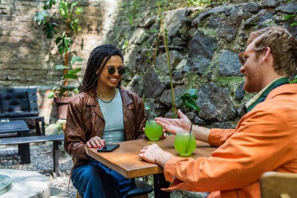 cheerful man pointing at happy african american friend in sunglasses near cocktail 