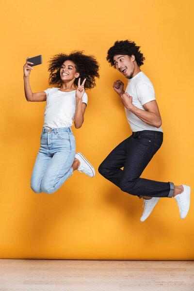 Portrait of a smiling young afro american couple taking a selfie while jumping isolated over yellow background