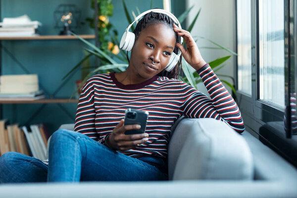 Shot of beautiful woman listening to music while using smartphone sitting on sofa at home.