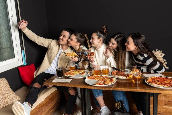 groups of young people take a selfie photo while eating pizzas at a restaurant