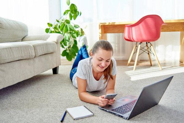 Portrait of happy smiling young woman lying on floor with laptop and using mobile phone at home