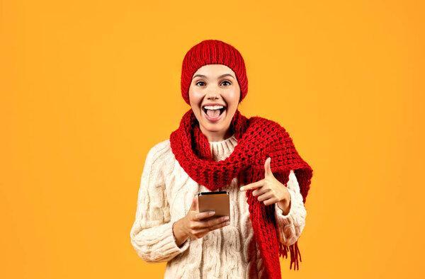 Winter Mobile Offer. Excited Young Woman Texting On Smartphone, Wearing Knitted Hat And Scarf, Advertising Application, Pointing Finger At Phone On Yellow Studio Background. Technology, Gadgets