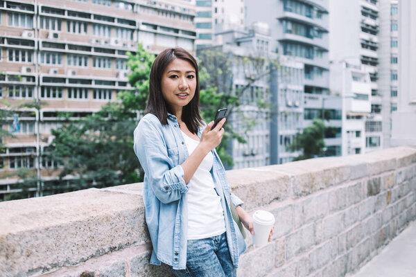 Lovely smiling ethnic female in casual clothes standing near brick fence and looking at camera while holding coffee and using telephone