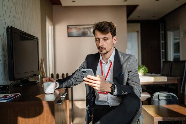 One man caucasian male adult businessman sitting at hotel room while take a brake from work use smarphone mobile phone for text messages or browse internet online hold cup of coffee in pause happy