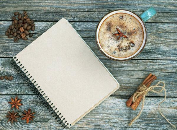 Mug of spice latter coffee, winter and autumn hot drink on wooden background. Blank craft notebook with cup of coffee, anise, cinnamon, beans of coffee. Flat lay. Fall mockup