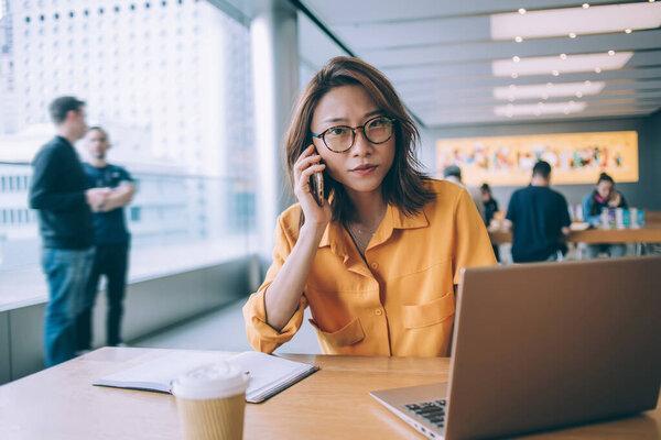 在香港大学的餐厅里，一位穿着黄色衬衫、戴着眼镜的有魅力的亚洲女学生在桌子旁打电话，桌上放着笔记本电脑和咖啡，并且看着镜头。