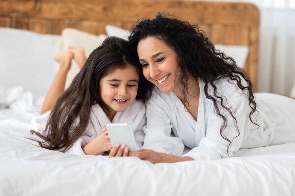 Cheerful mother and daughter in bathrobes using smartphone