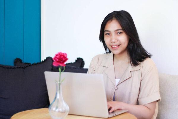 happy asian woman sit on cozy couch using laptop to have online business meeting, freelance project, wearing casual fashionable clothes, looking at camera