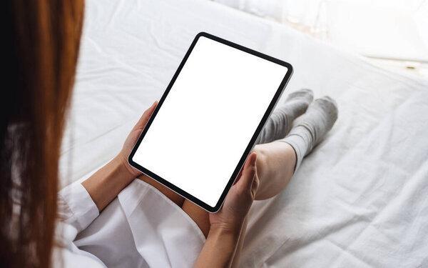Mockup image of a woman holding black tablet pc with blank desktop white screen while sitting on a cozy white bed at home