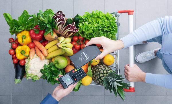 Woman pushing a full shopping cart and paying for groceries with her smartphone, mobile payments concept, top view
