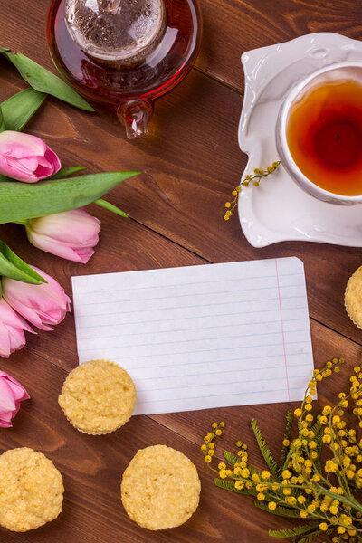 Women's day greeting card with tulips, mimosa, tea and cupcakes on brown wooden background.