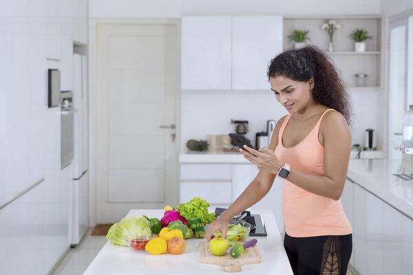 Young woman using a smartphone and cooking vegetables in the kitchen