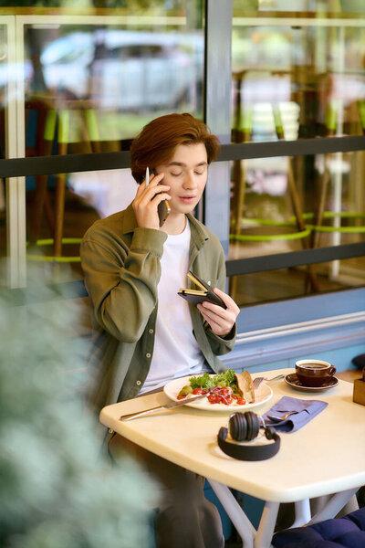 A young man in an olive green shirt savors his meal while talking on the phone in a modern cafe.