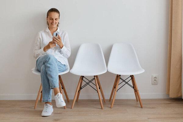 Indoor shot of young adult Caucasian smiling woman wearing white shirt and jeans sitting on chair against wall, using cell phone while waiting her queue.