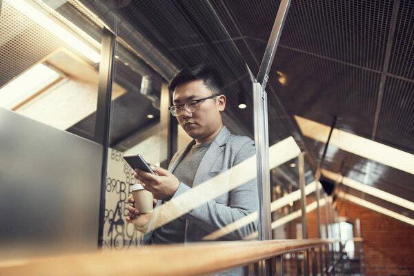 Waist up portrait of young Asian businessman using smartphone standing behind glass wall in modern office interior, copy space