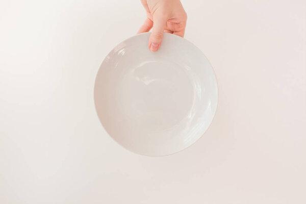  Female hand holds a white round plate on a white background as a mockup with copy space. Ceramic flat saucer.