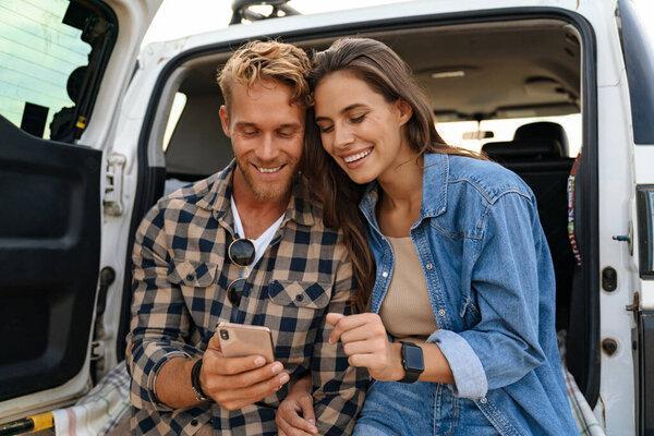 Beautiful couple sitting at the back of car and using smart phones