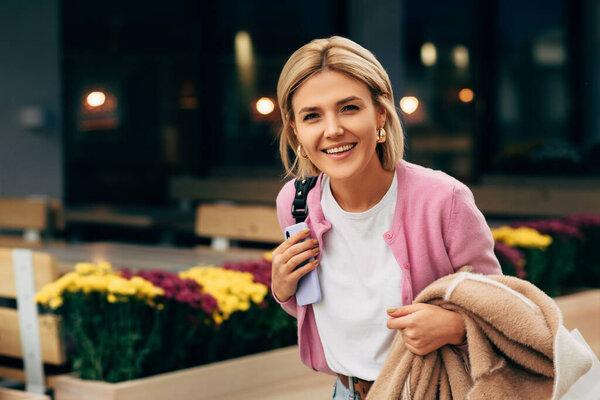 Happy gorgeous blonde young woman wearing white t-shirt and pink jacket, smiling and walking on the city street. Pretty female student posing in the street.