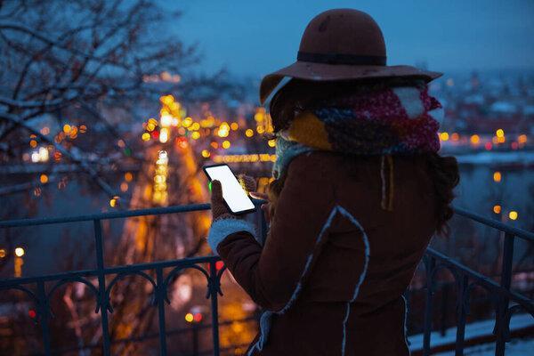 Seen from behind middle aged woman in brown hat and scarf outside in the city park in winter in sheepskin coat using smartphone applications at night.