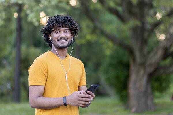 Portrait of a smiling Indian man doing sports and jogging in the park. Standing in headphones, holding a phone and smiling at the camera. Close-up photo.