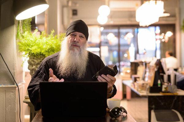 Mature bearded hipster man using laptop and looking confused at the coffee shop