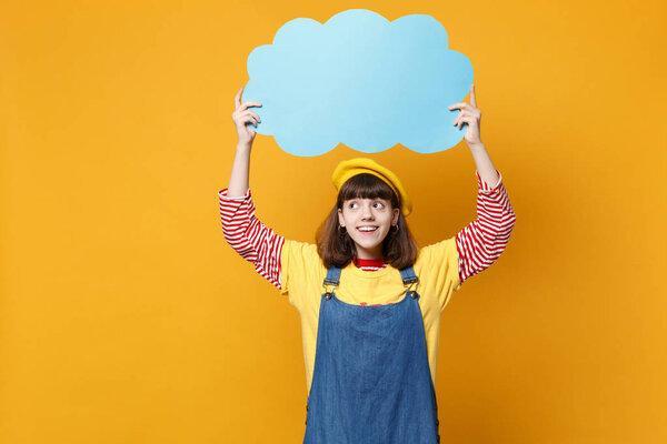 Cheerful girl teenager in french beret, denim sundress hold blue empty blank Say cloud, speech bubble isolated on yellow wall background. People sincere emotions lifestyle concept. Mock up copy space