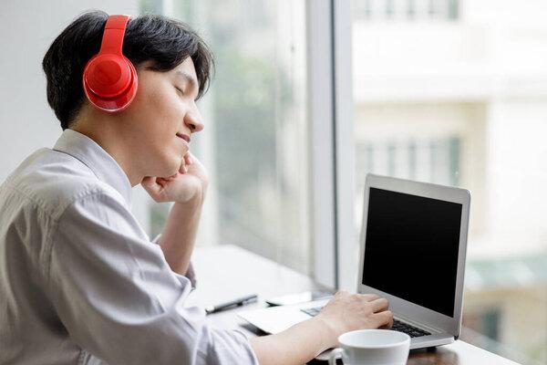 young asian businessman sit and drinks coffee by window in office wear headphone using laptop for listen to music,