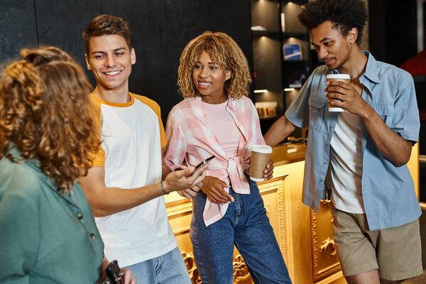 handsome young man with mobile phone near smiling multiethnic buddies in lobby of modern hostel