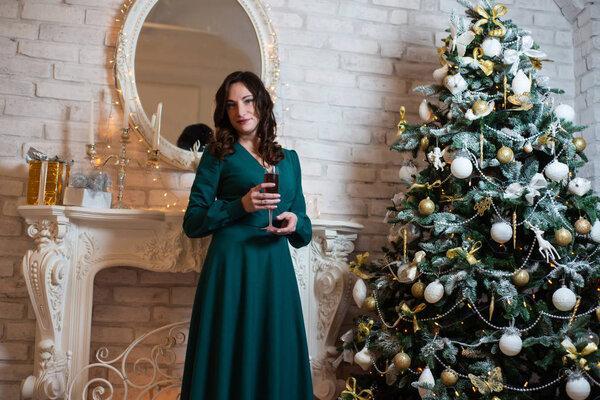 Beautiful woman in green dress posing near the Christmas tree and fireplace