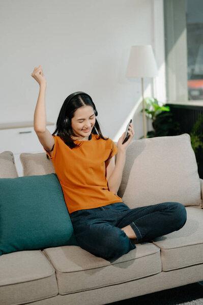 Happy Young asian woman using smartphone and headphones while seated on couch at home 