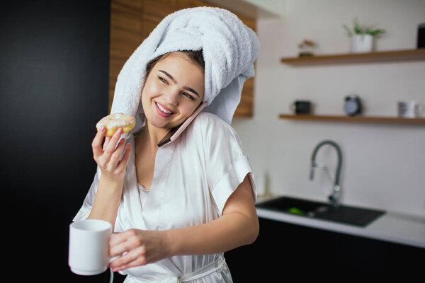 Young woman in kitchen during quarantine. Talking on phone and smile. Breakfast after bathing. Woman in white robe and towel eat donut.