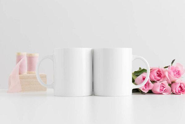 Two mugs mockup with a bouquet of pink roses and silk ribbons on a white table.