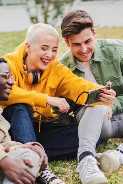 Vertical portrait of a group of teenage students having fun using a cellphone to browse on internet. Young people smiling and laughing watching a smartphone social media app sitting at meadow grass