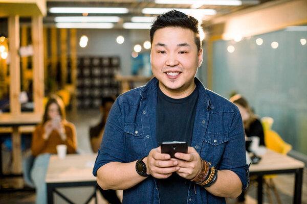 A young Chinese businessman in casual wear smiling and looking at camera, holding smartphone, standing in coworking room, friends on the background